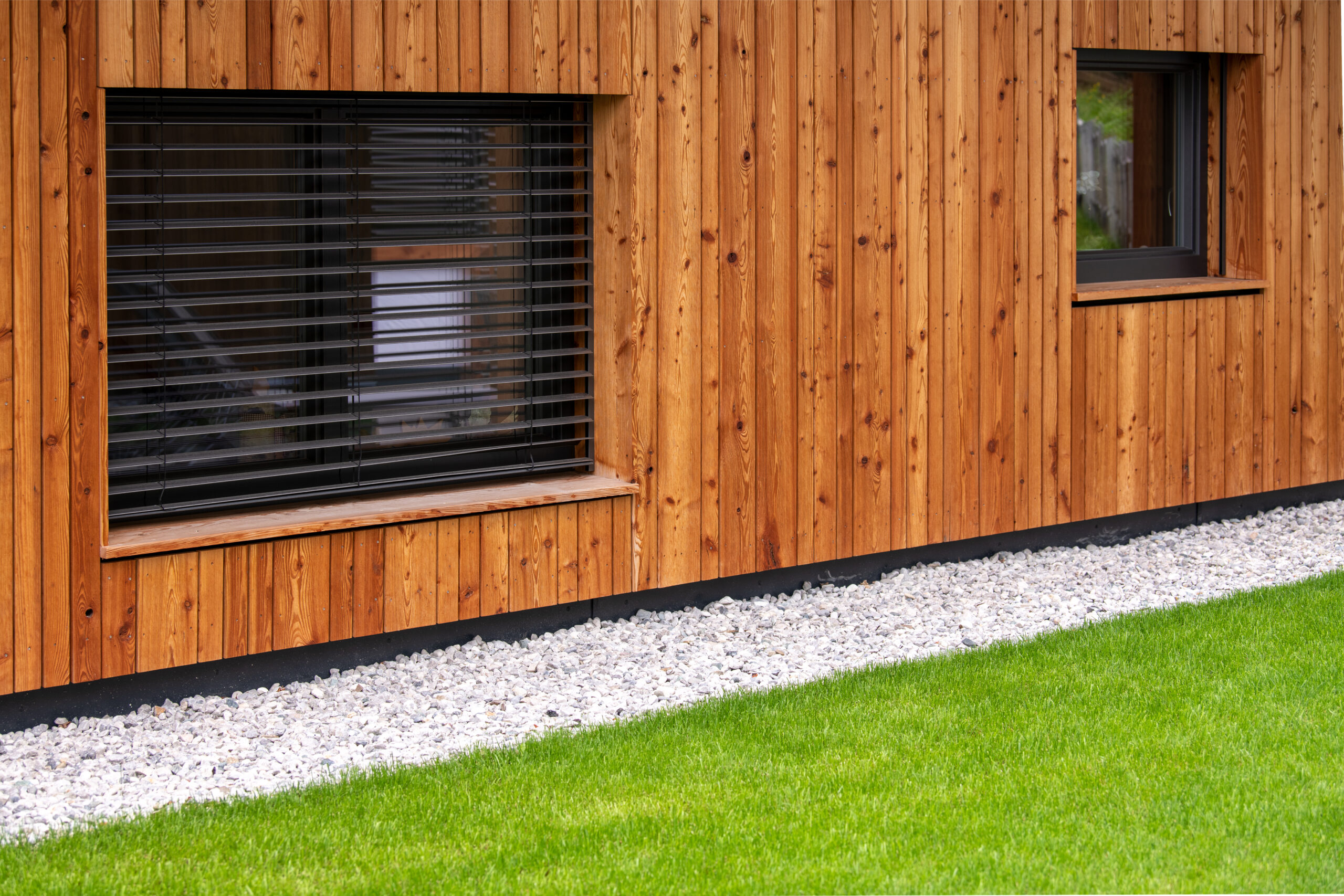 Moderne Architektur mit natürlichen Materialen- Fassade aus Holz, davor sehr gepflegter grüner Rasen und Kiesstreifen - moderne Fenster mit Raffstores